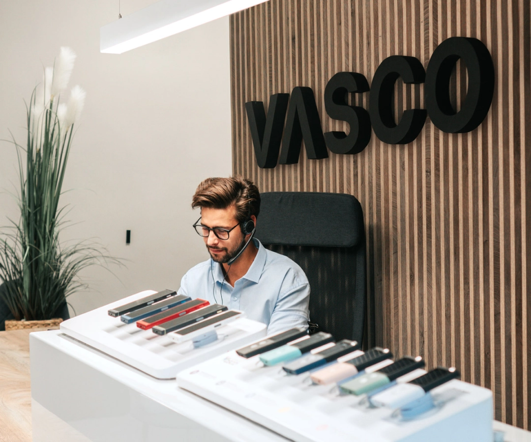 Een man van de klantenservice zit aan een bureau met meerdere Vasco Translator-apparaten in verschillende kleuren, met een groot 'VASCO'-bord op de houten muur achter hem.
