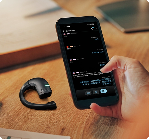 Close-up van een hand die een smartphone vasthoudt waarop een realtime vertaalchat tussen het Engels en het Chinees te zien is. Naast de telefoon ligt een zwarte Vasco-vertaaloorplug met een groen oplichtend LED-lampje op een houten bureau. 