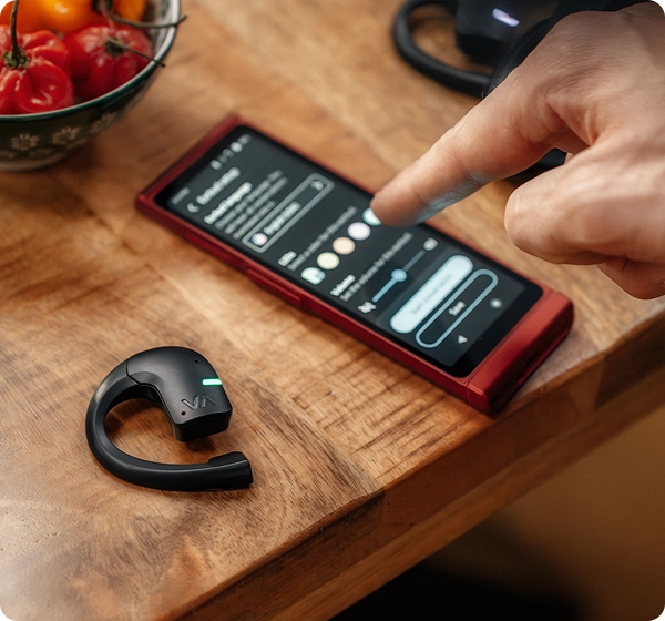 Een close-up van iemands vinger die instellingen zoals kleur en volume aanpast op het scherm van een rood Vasco Translator V4-apparaat. Ernaast op de houten tafel ligt een zwarte Vasco Translator E1-oortelefoon met een groen oplichtend LED-lampje, terwijl op de achtergrond een schaal met rode paprika’s te zien is. 