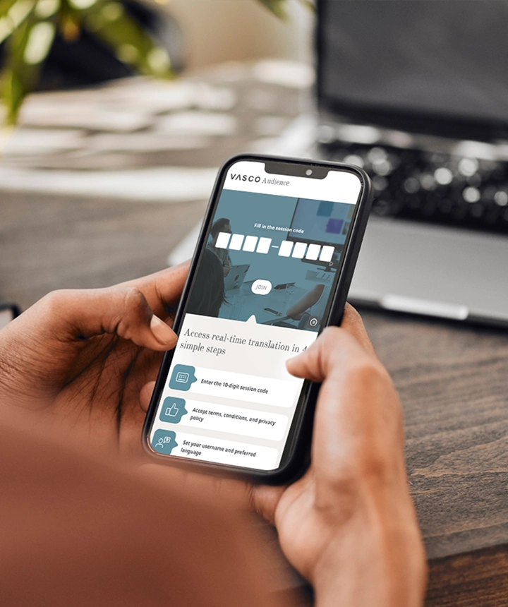 Een blik over de schouder van iemand die een smartphone vasthoudt waarop de webinterface van Vasco Audience te zien is. Op het scherm staat een invoerveld met de tekst 'Vul de sessiecode in' en een witte knop met de tekst 'DEELNEMEN', boven een lijst met instructies met de tekst 'Toegang tot realtime vertaling in 4 eenvoudige stappen'. Op de achtergrond zijn een wazig houten bureau en een laptop te zien.