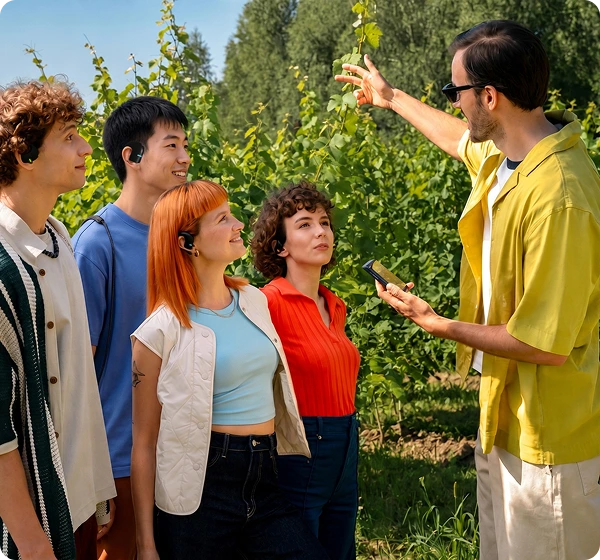 Een groep van vijf mensen buiten in een zonovergoten wijngaard. Rechts houdt een man in een geel shirt en met een zonnebril op een Vasco Q1-vertaalapparaat vast en wijst naar de groene wijnstokken. Links luisteren vier mensen aandachtig; ze dragen allemaal een zwarte Vasco E1-vertaalapparaat, waarmee de functie voor realtime groepsgesprekken wordt gedemonstreerd. 