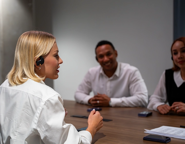 Een profielfoto van een blonde vrouw in een witte blouse die aan het woord is tijdens een zakelijke vergadering aan een houten vergadertafel. Ze draagt een zwarte Vasco Translation E1-oortelefoon. Op de wazige achtergrond zitten twee andere zakenmensen aan de andere kant van de tafel; ze glimlachen en luisteren aandachtig. 