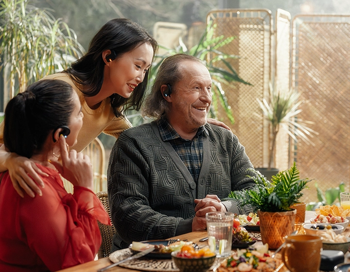 Een hartverwarmende familiebijeenkomst met meerdere generaties rond een eettafel vol eten. Een oudere, glimlachende man in een grijs vest en twee vrouwen maken vrolijk een praatje in een zonovergoten kamer vol planten. Alle drie dragen ze zwarte Vasco Translation E1-oordopjes. 