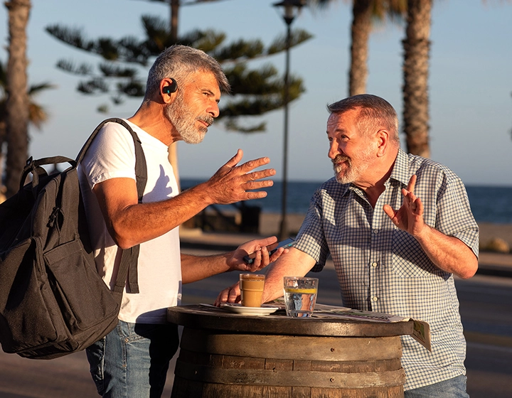 Twee mannen staan bij een tafel buiten en voeren een gesprek. Een van hen draagt een rugzak en beiden gebruiken Vasco Translator E1-oordopjes. 