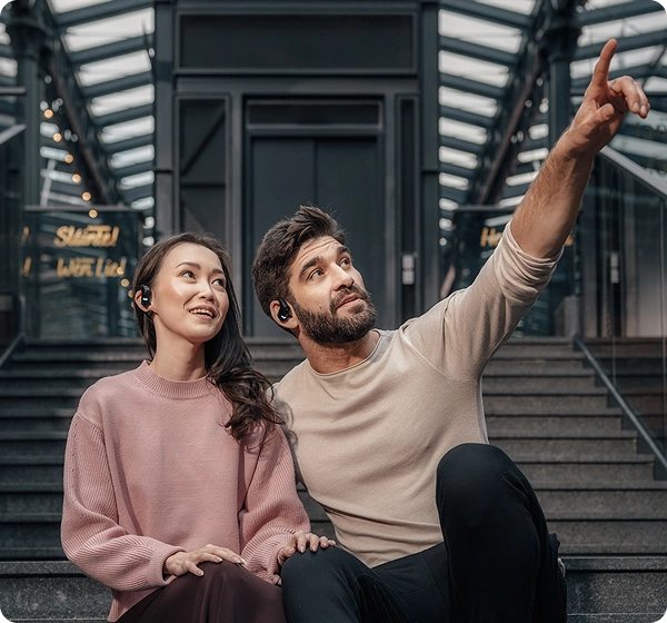 Een man en een vrouw zitten op betonnen treden in een modern gebouw met een glazen overkapping. De man wijst met zijn hand omhoog, terwijl beiden zwarte Vasco Translation E1-oordopjes dragen en in de richting van zijn gebaar kijken. 