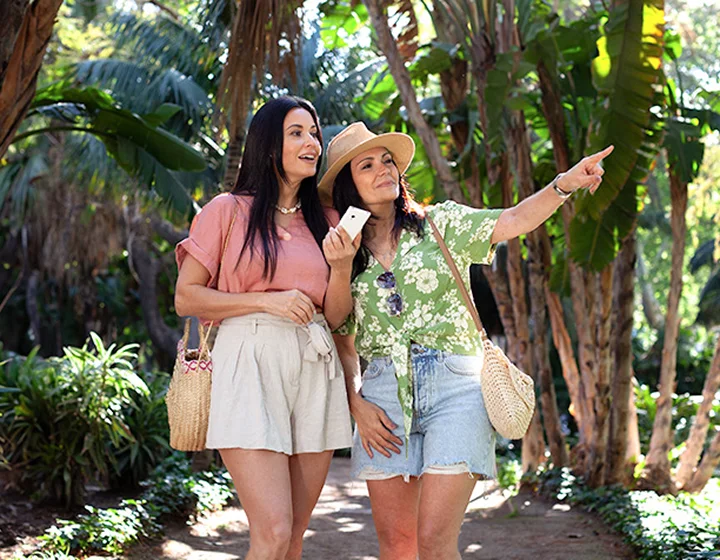 Twee vrouwen in een vakantieomgeving kijken in de richting waar een van hen naar wijst. Een van de vrouwen houdt een Vasco Translator V4 in haar hand.