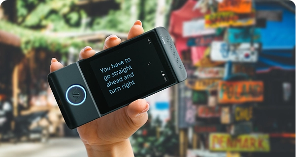 Een close-up van een hand die een zwarte elektronische vertaler van Vasco horizontaal vasthoudt. Het scherm staat in volledig scherm, waarop de vertaalde tekst in een groot, helder blauw lettertype wordt weergegeven: 