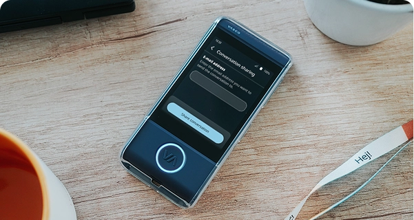 Een close-up vanuit een hoge hoek van een donkerblauwe elektronische vertaler van Vasco in een doorzichtig beschermhoesje, die op een lichtgekleurde houten tafel ligt. Het scherm van het apparaat is actief en toont een menu 'Gesprek delen' met een veld om een e-mailadres in te voeren en een knop met de tekst 'Gesprek delen'. Rondom het apparaat op de tafel liggen de rand van een oranje koffiemok, een witte plantenpot en een wit lanyard met het woord 'Hej!' erop gedrukt. De scène is helder verlicht, wat een strakke en moderne uitstraling creëert. 