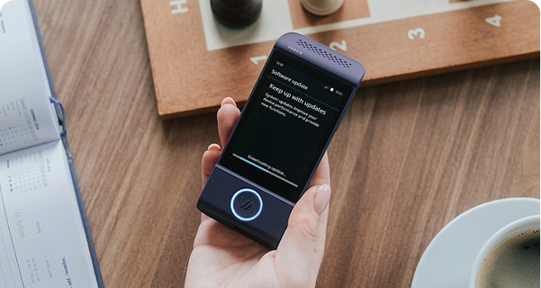 Een opname van bovenaf van een hand die een paarse elektronische vertaler van Vasco boven een houten tafel houdt. Op het scherm van het apparaat staat het bericht „Software-update“ met de tekst „Blijf op de hoogte van updates“ en een voortgangsbalk met de tekst „Update wordt gedownload...“. Op de tafel rondom het apparaat liggen een houten schaakbord met schaakstukken, een open kalender en een wit kopje koffie. De verlichting is zacht en natuurlijk, wat een ontspannen, alledaagse sfeer creëert. 