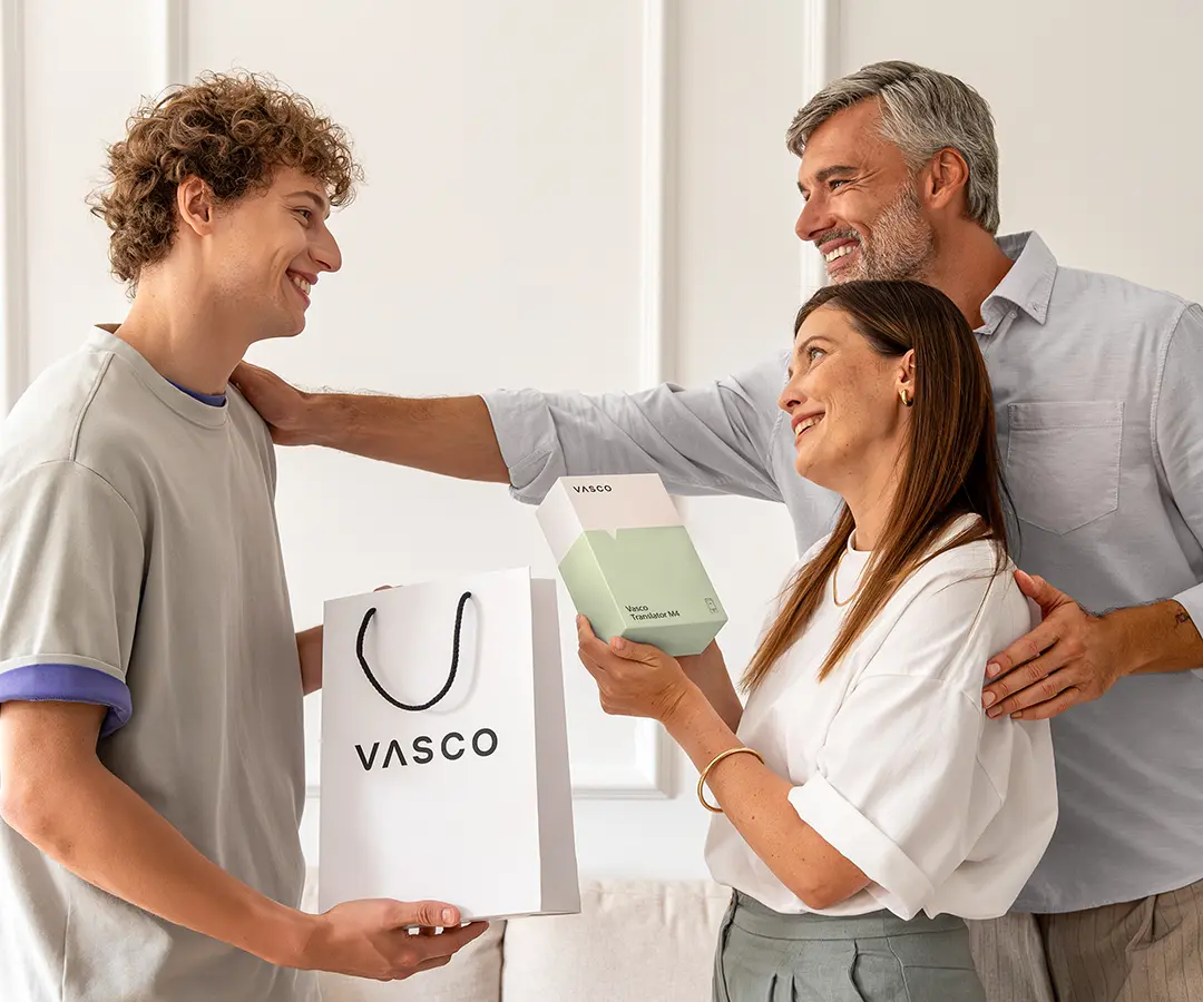 Een hartverwarmend tafereel van drie mensen die glimlachend bij elkaar staan. Links houdt een jonge man met krullend haar een witte „VASCO“-boodschappentas vast. In het midden houdt een vrouw een wit- en lichtgroene doos met het opschrift „Vasco Translator M4“ vast, terwijl ze de jonge man blij aankijkt. Achter haar staat een man met grijzend haar die warm glimlacht, met één hand op de schouder van de vrouw en de andere op de schouder van de jonge man. Ze bevinden zich in een lichte kamer met een neutraal gekleurde muur en een bank op de achtergrond. 