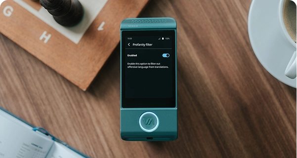 Een bovenaanzicht van een blauwgroene elektronische vertaler van Vasco die op een houten tafel ligt. Het scherm van het apparaat is actief en toont het instellingenmenu voor het „Scheldwoordfilter“, waarbij de schakelaar op „Ingeschakeld“ staat. Onder de schakelaar staat de volgende beschrijving: „Schakel deze optie in om aanstootgevend taalgebruik uit vertalingen te filteren.“ Het apparaat is omringd door lifestyle-items, waaronder een houten schaakbord met een donkere schaakstuk, een hoek van een open notitieboekje en een witte koffiekop, allemaal vastgelegd in zacht, natuurlijk licht. 
