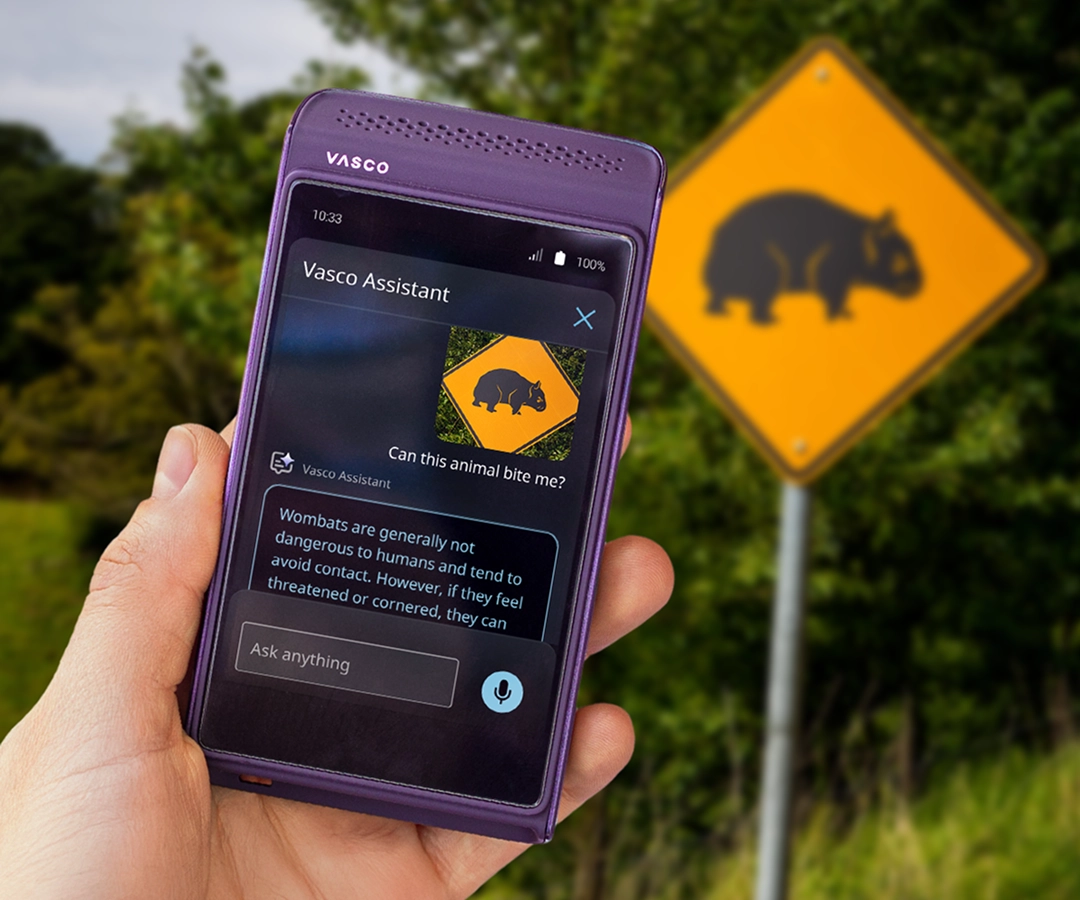 Een hand houdt een paarse Vasco elektronische vertaler vast. De Vasco Assistant-app is zichtbaar op het scherm van het apparaat en toont een foto van een waarschuwingsbord met een afbeelding van een wombat en een beschrijving van deze dieren. Op de achtergrond, achter de hand die het apparaat vasthoudt, is een weg te zien met hetzelfde gele verkeersbord met een wombat, dat ook op het scherm van de vertaler verschijnt.