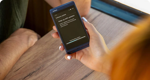 Een opname over de schouder van een vrouw die in een trein een blauwe Vasco Translator Q1 boven een houten tafel houdt. Op het scherm van het apparaat is een melding ‘Software-update’ te zien, met een voortgangsbalk die aangeeft: ‘Update wordt gedownload...’.